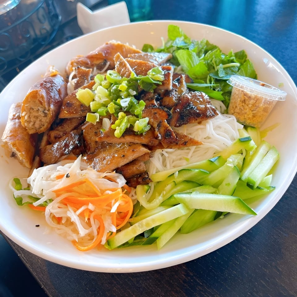 A large bowl of Vietnamese vermicelli noodles with grilled pork, spring rolls, and fresh vegetables.