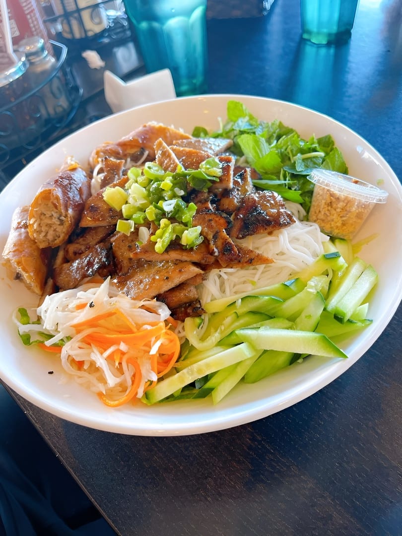 A large bowl of Vietnamese vermicelli noodles with grilled pork, spring rolls, and fresh vegetables.