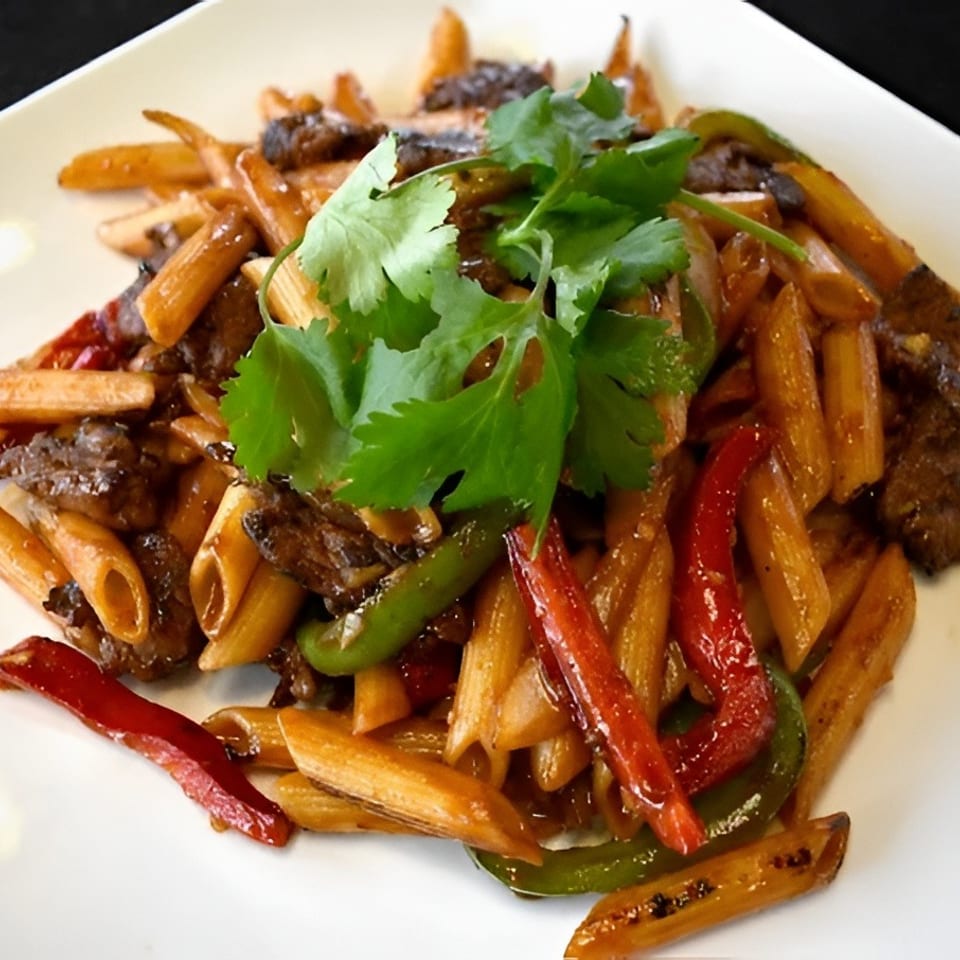 Penne pasta with beef, red and green bell peppers, and cilantro on a white plate.