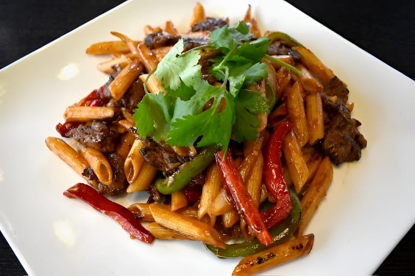 Penne pasta with beef, red and green bell peppers, and cilantro on a white plate.