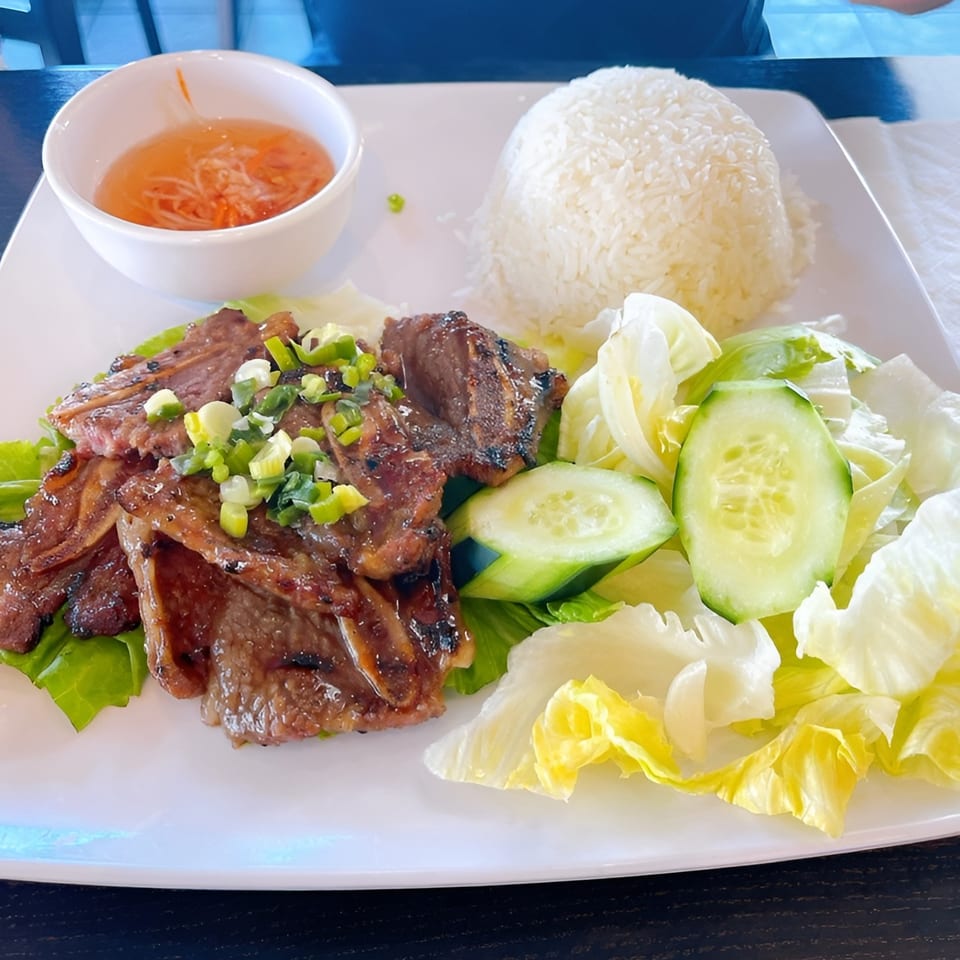 Grilled short ribs with rice, salad, and dipping sauce on a white plate.