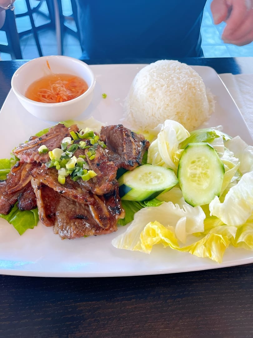 Grilled short ribs with rice, salad, and dipping sauce on a white plate.